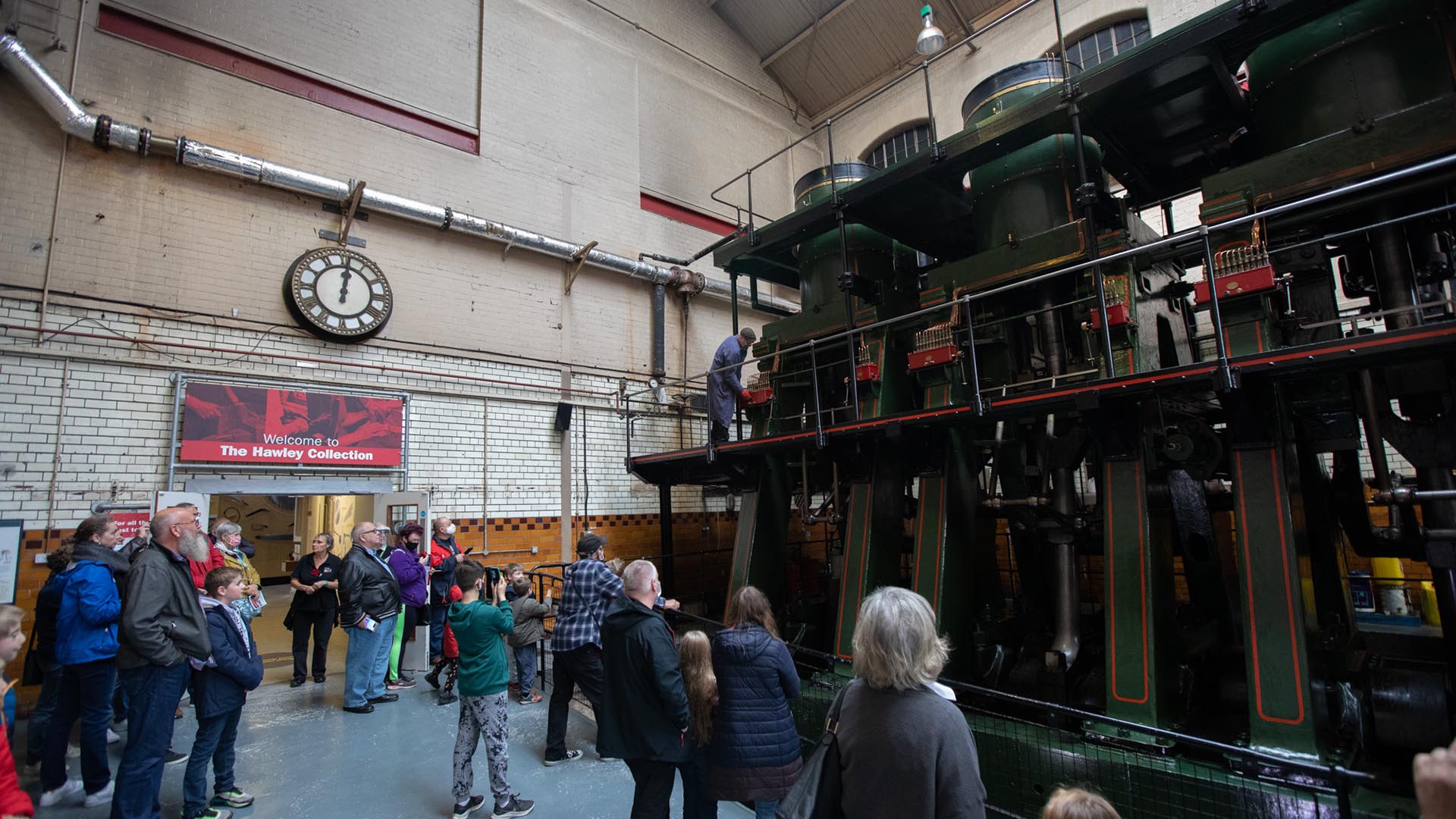 A group of visitors at Kelham Island Museum stand behind a metal barrier to watch an engineer run the River Don engine. The large steam engine is dark green, red, black and gold. The mechanics are all visible. It has exposed pistons, three cylinders at the top and two platforms around it.